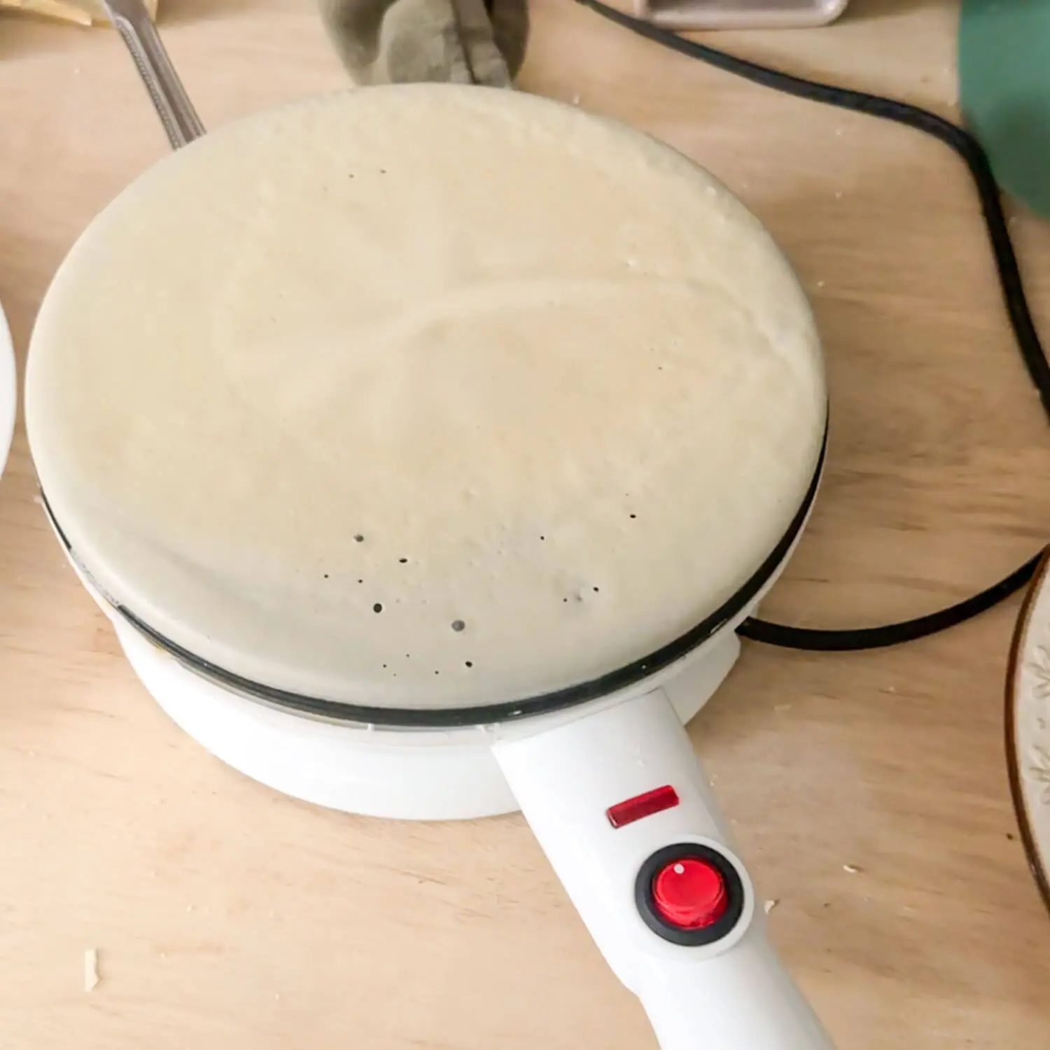 Cordless crepe maker with batter coating the nonstick plate, red power light on, showing the dip and flip step for fast, thin crepes at home.