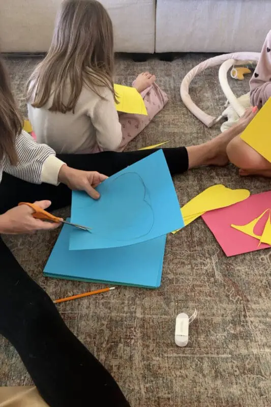 Cutting a large petal shape from blue cardstock for a no-sew flower costume, kids crafting nearby with yellow and pink pieces on the floor.