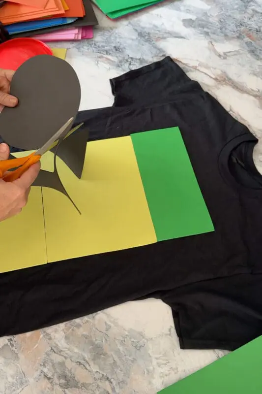Hands cutting a black foam circle to create a logo for a DIY costume t-shirt, part of an easy and funny Halloween craft project.