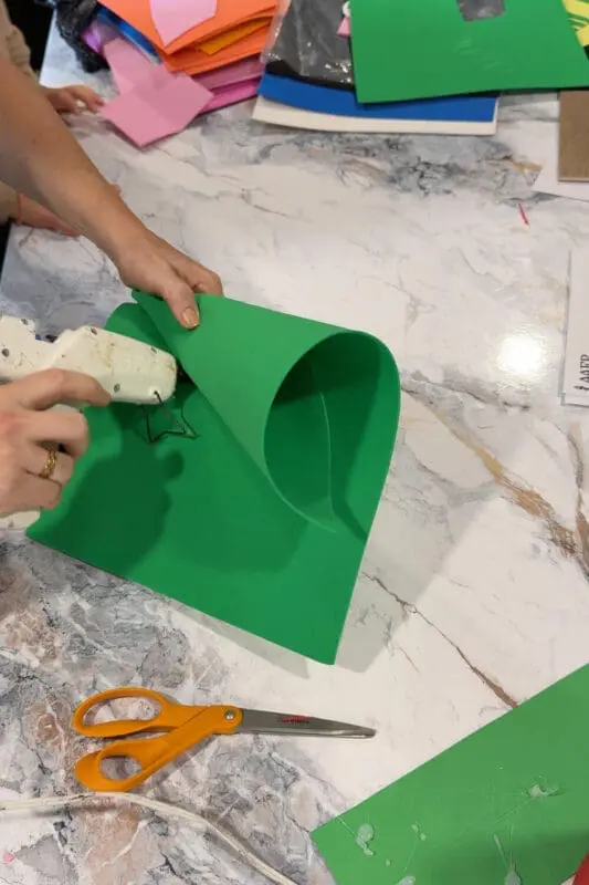 Gluing green foam sheet into a cone shape using a hot glue gun to create a garden gnome hat for a DIY kids costume.