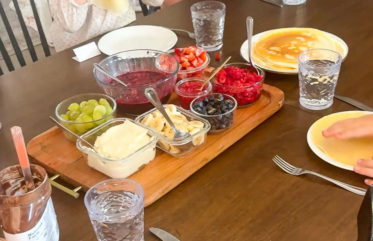 Family breakfast with a stack of homemade crepes and toppings—berries, bananas, grapes, berry compote, whipped cream, and Nutella—kids assembling their own breakfast.