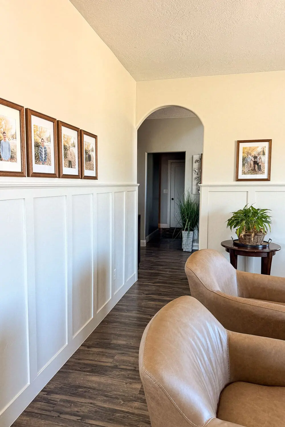 Formal living room wall with white board and batten, arched doorway, family photo gallery, wood floors, and tan leather chairs in the foreground.