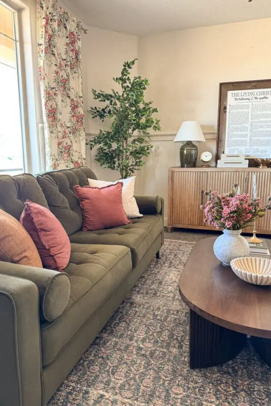 Formal living room with floral curtains, green velvet sofa, pink pillows, faux tree, ribbed wood credenza, and styled coffee table showing how brighter drapery lightens the space.