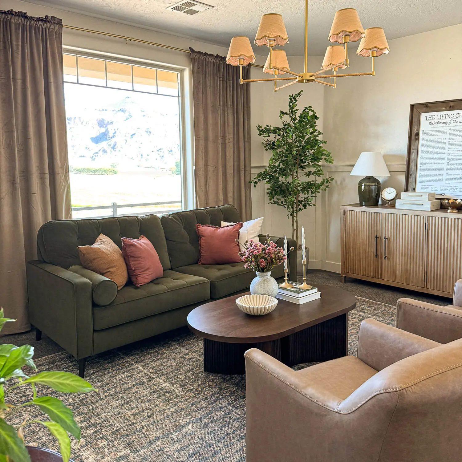 Formal living room makeover with olive green sofa, tan leather chairs, oval wood coffee table, brass chandelier with scalloped shades, and taupe curtains framing a large window.