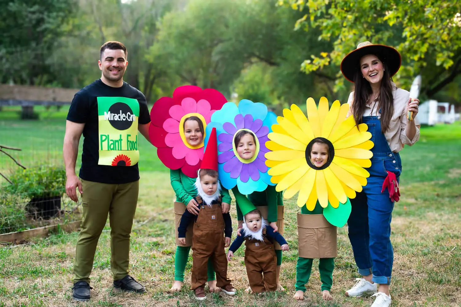 Family garden-themed Halloween costumes—three kids in no-sew cardstock flower costumes with flower-pot skirts (red, blue-purple, yellow sunflower), two toddlers as garden gnomes, dad in a Miracle-Gro plant food shirt, and mom dressed as a gardener with a straw hat and trowel.