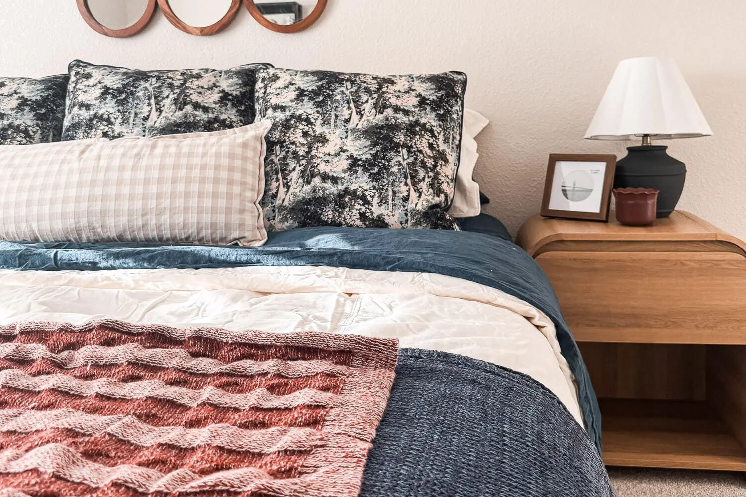 close up of cozy fall bedding layered with navy quilt, cream duvet, rust knit throw, beige gingham lumbar, patterned euro pillows, and a wood nightstand with lamp and art in a guest room refresh