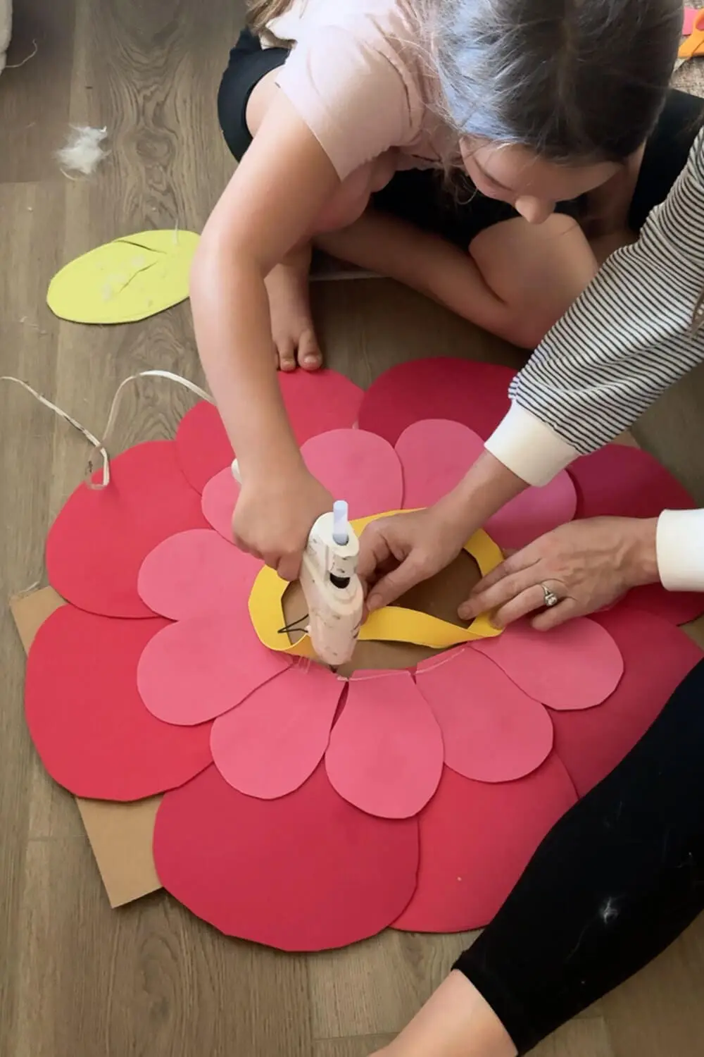 Child using a hot glue gun to attach pink and red cardstock petals around a yellow face ring on a cardboard base for a no sew flower costume.