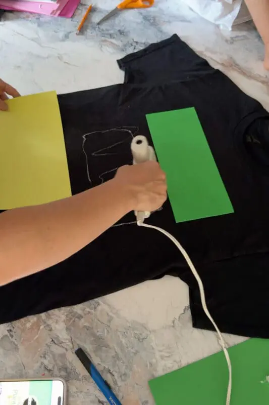 Hands using a hot glue gun to attach colorful foam panels to a black shirt while assembling a DIY costume t-shirt for a funny Halloween craft project.