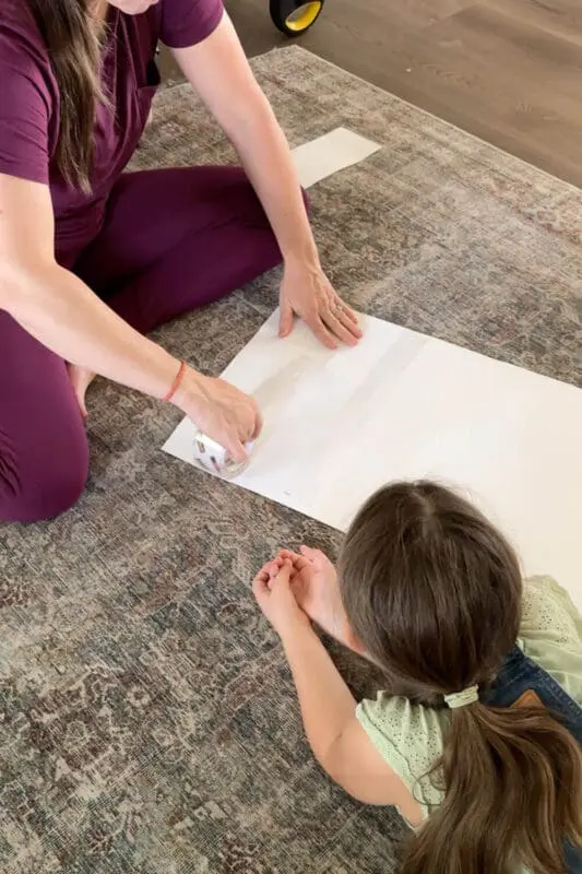 Taping white poster board into a long strip on the floor to form the base of a flower-pot skirt for a no-sew cardstock flower costume.