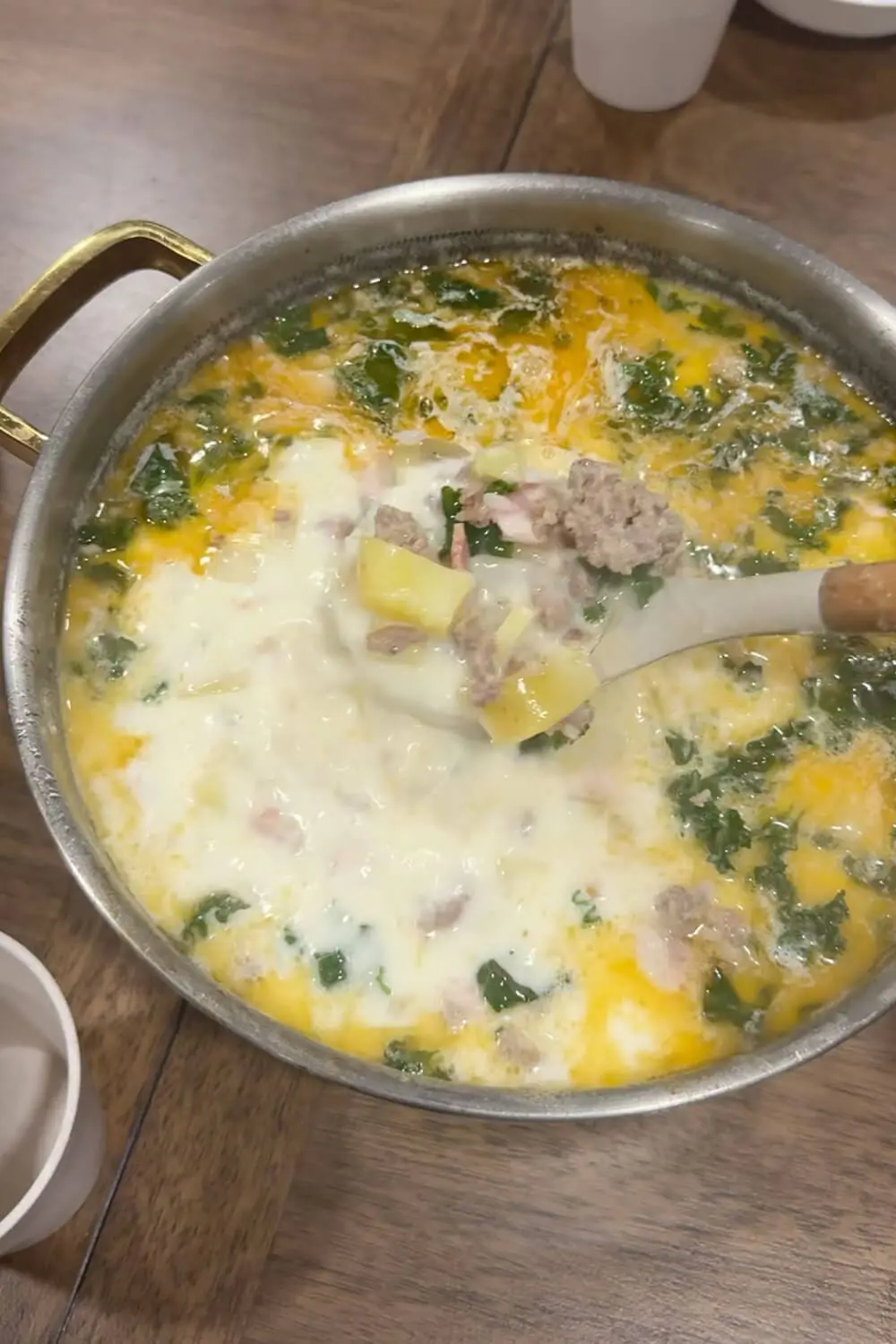 Overhead view of creamy Zuppa Toscana in a stainless pot, with a ladle scooping Yukon gold potatoes, Italian sausage, bacon, and kale in a garlic-cream broth.