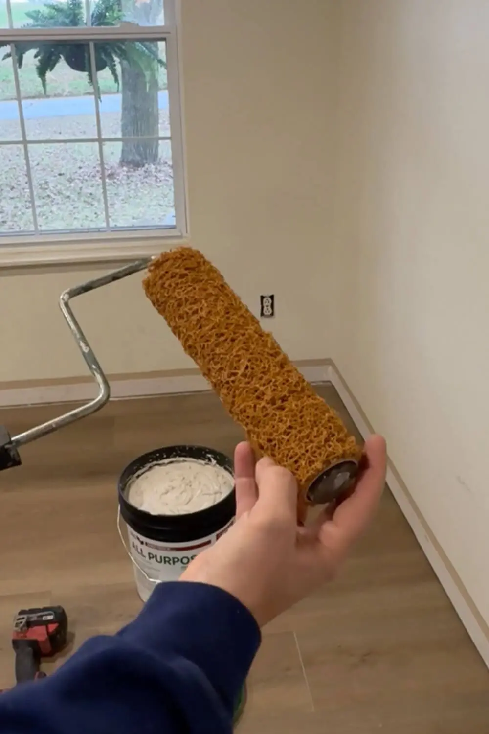 Close-up of the nap roller used for skim coating a wall, held next to a bucket of premixed joint compound before applying