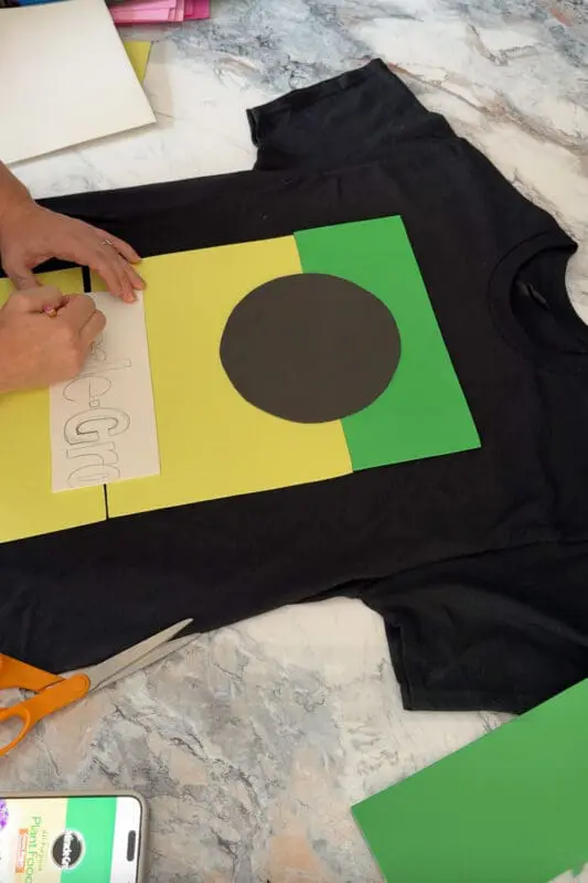 Tracing letters onto white craft foam beside a black, yellow, and green layout while creating a DIY costume t-shirt design for an easy, funny Halloween project.
