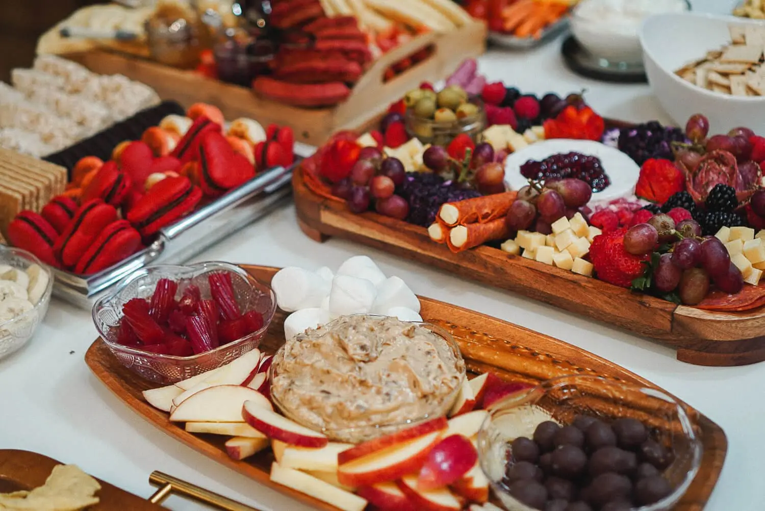 Apple dip tray with sliced apples, marshmallows, and pretzels in the foreground, set among assorted charcuterie and dessert boards on a party table.