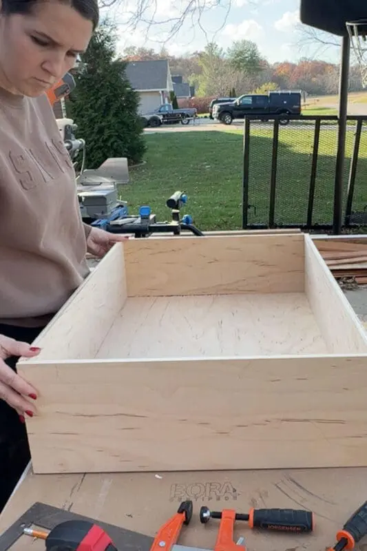 Woman checking the fit of a newly assembled plywood drawer box on an outdoor workbench.