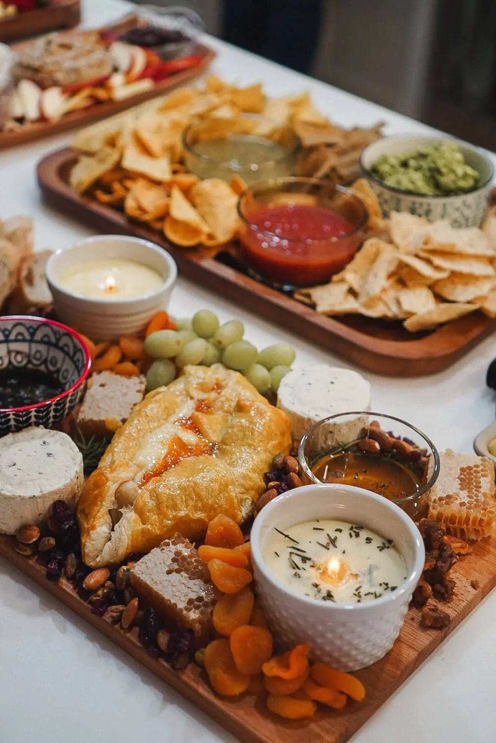 Baked brie charcuterie board with honeycomb, nuts, dried apricots, and warm rosemary brie; chips and dips tray behind it with salsa, salsa verde, and guacamole.