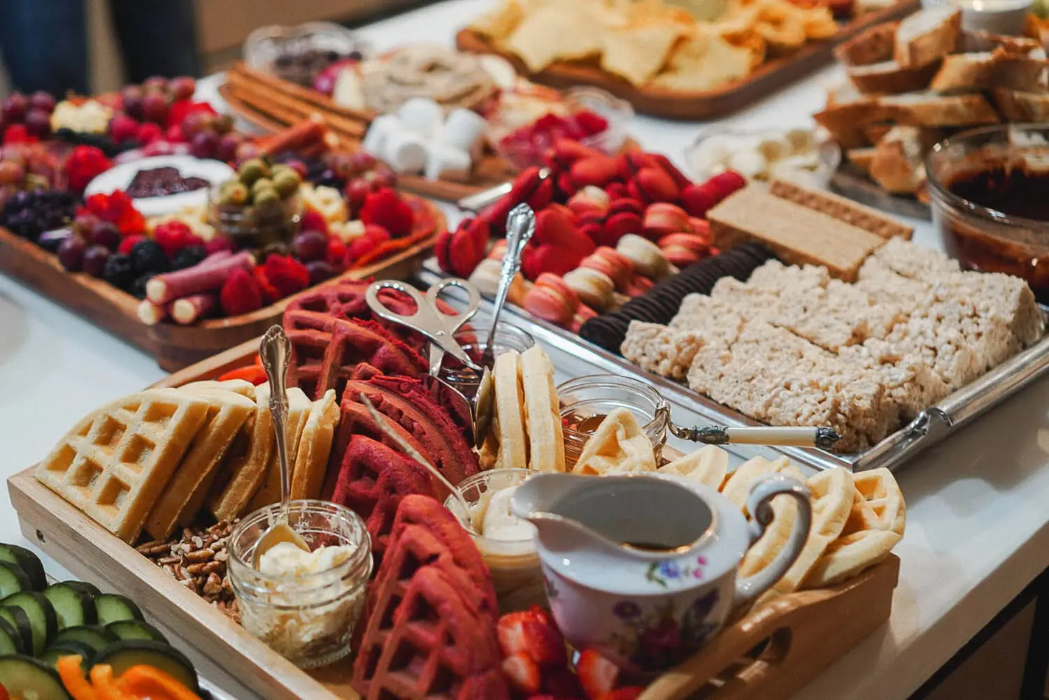 Brunch charcuterie board with red-velvet and classic waffles, butter and syrup, strawberries and pecans, plus a dessert spread in the background with macarons, Oreos, and Rice Krispies treats—fun charcuterie board ideas for a party.