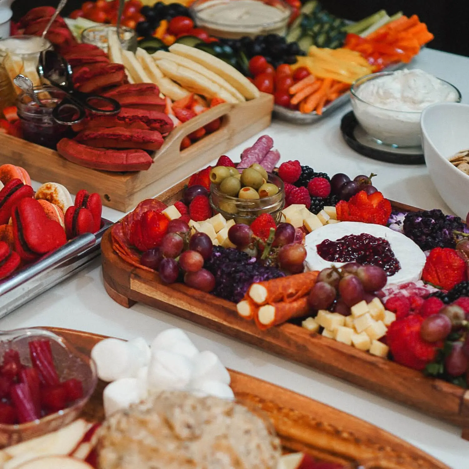 Charcuterie board ideas for a party—a table of boards with brie topped with jam, grapes, olives, salami, cheese cubes, red macarons, waffles, and a veggie tray with dips.