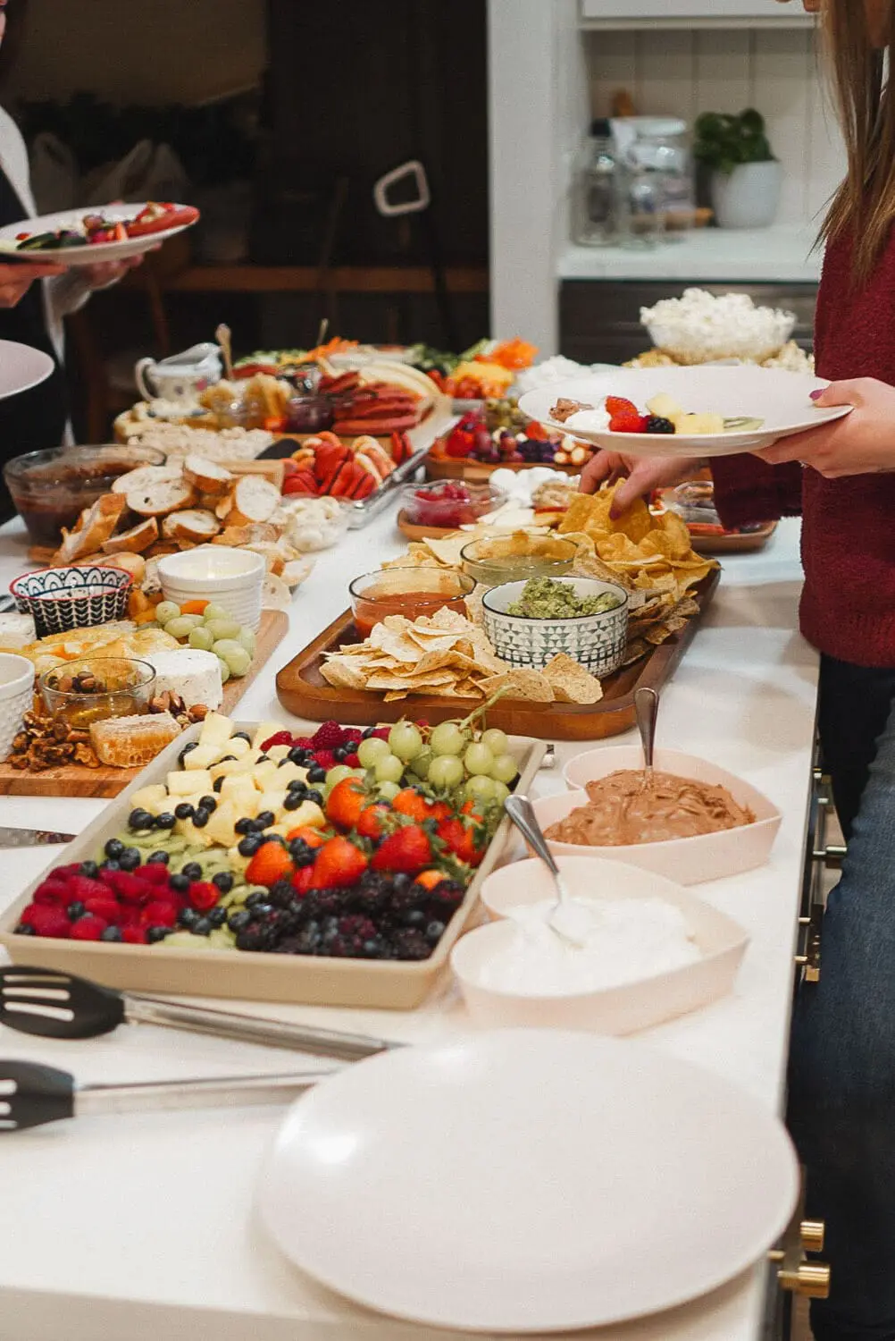 Guests serving from a charcuterie buffet—fruit tray with chocolate and marshmallow dips, chips with guacamole and salsa, plus breads, cheese, and veggie boards in the background at a potluck charcuterie party.
