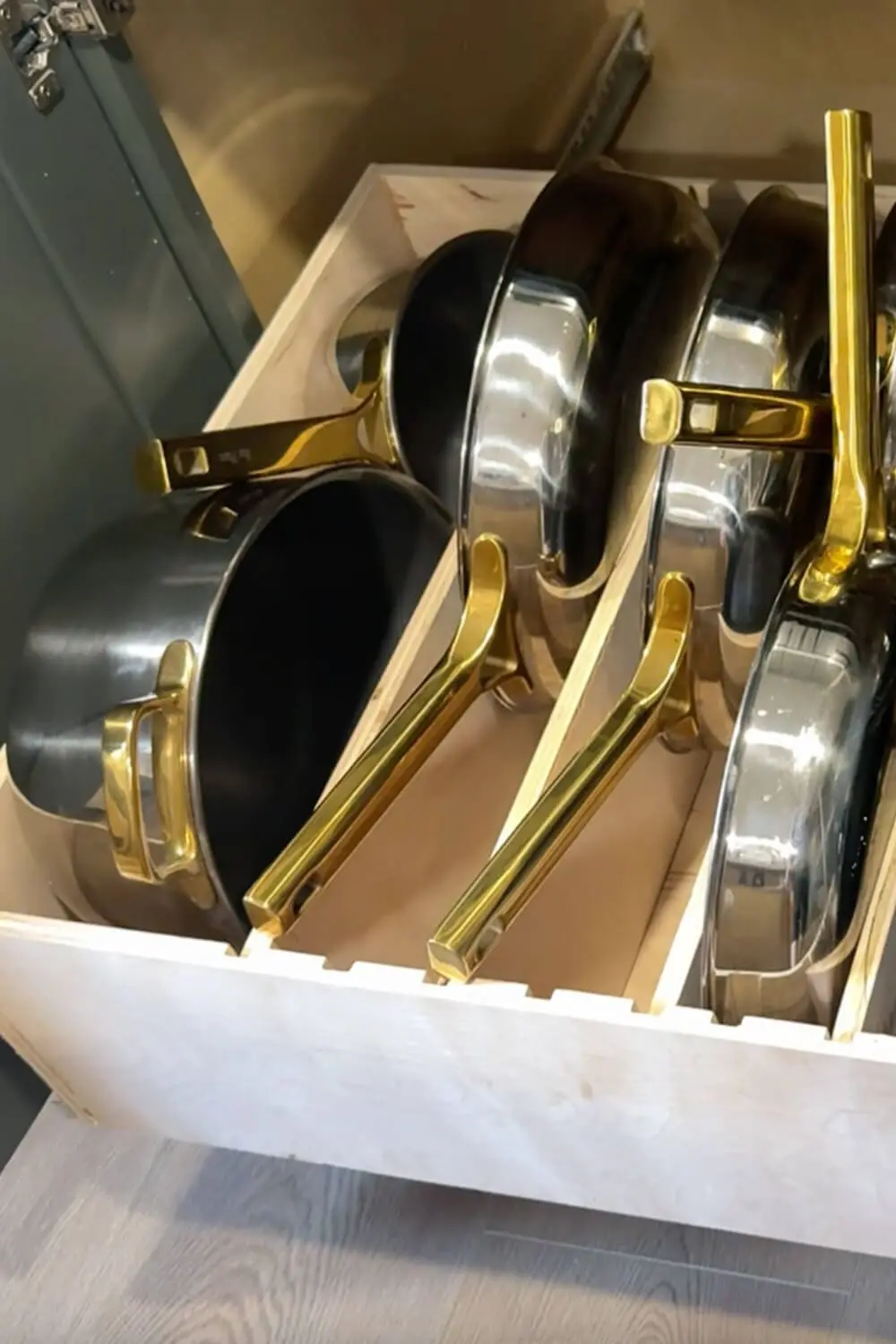 Close-up of stainless steel pots and pans with brass handles standing upright in vertical plywood dividers inside a DIY slide-out drawer, showing organized pots and pans storage in a lower cabinet.