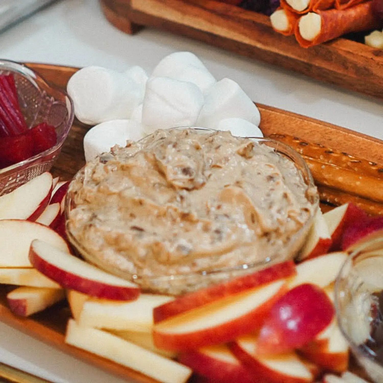 Close-up bowl of creamy brown sugar cream cheese apple dip with toffee bits, surrounded by red apple slices, marshmallows, and pretzel rods on a wooden tray.