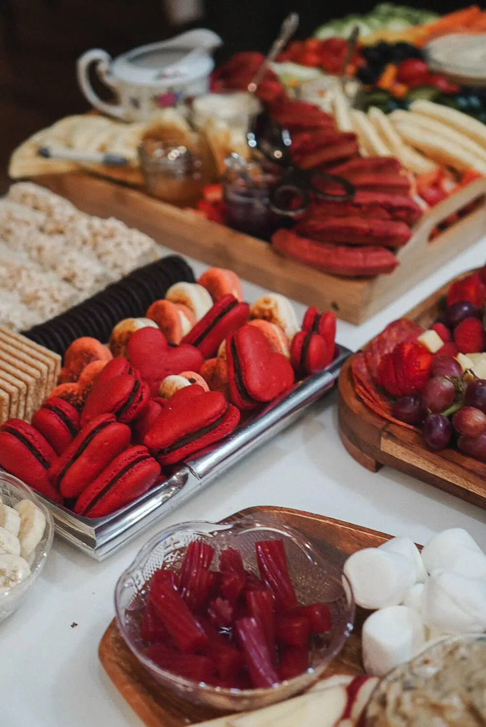 Dessert tray at a Galentine's Gathering with bright red heart macarons, Oreos, graham crackers, red licorice bites, and marshmallows, with fruit and waffles in the background.