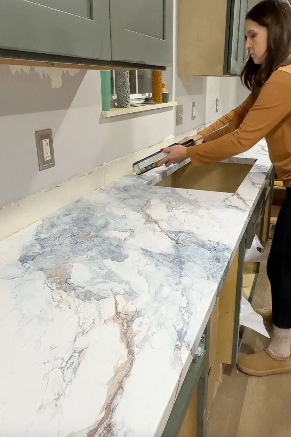 Woman installing marble contact paper on new DIY countertops, smoothing the faux marble finish across a long plywood kitchen counter.