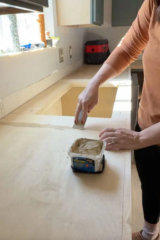 Woman spreading wood filler over a plywood kitchen countertop seam to prep DIY countertops for a faux marble epoxy finish.