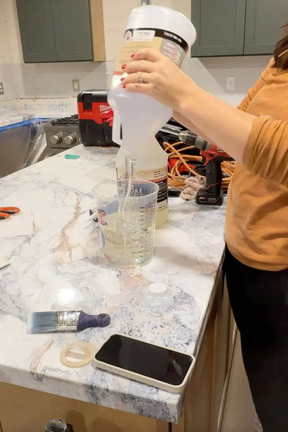 Close-up of DIY countertops that look like marble while epoxy resin is being poured into a mixing cup to seal and protect the faux marble finish.