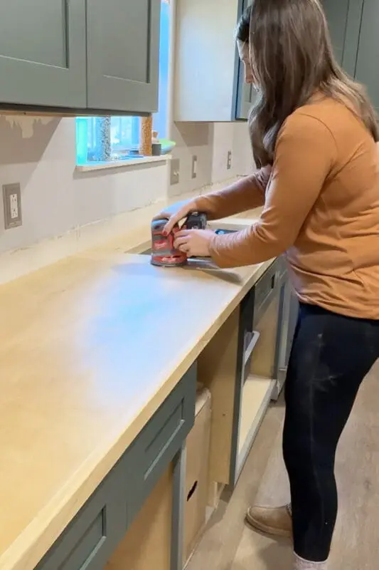 Woman sanding freshly installed plywood kitchen countertops smooth with a power sander while prepping DIY countertops for a marble epoxy finish.