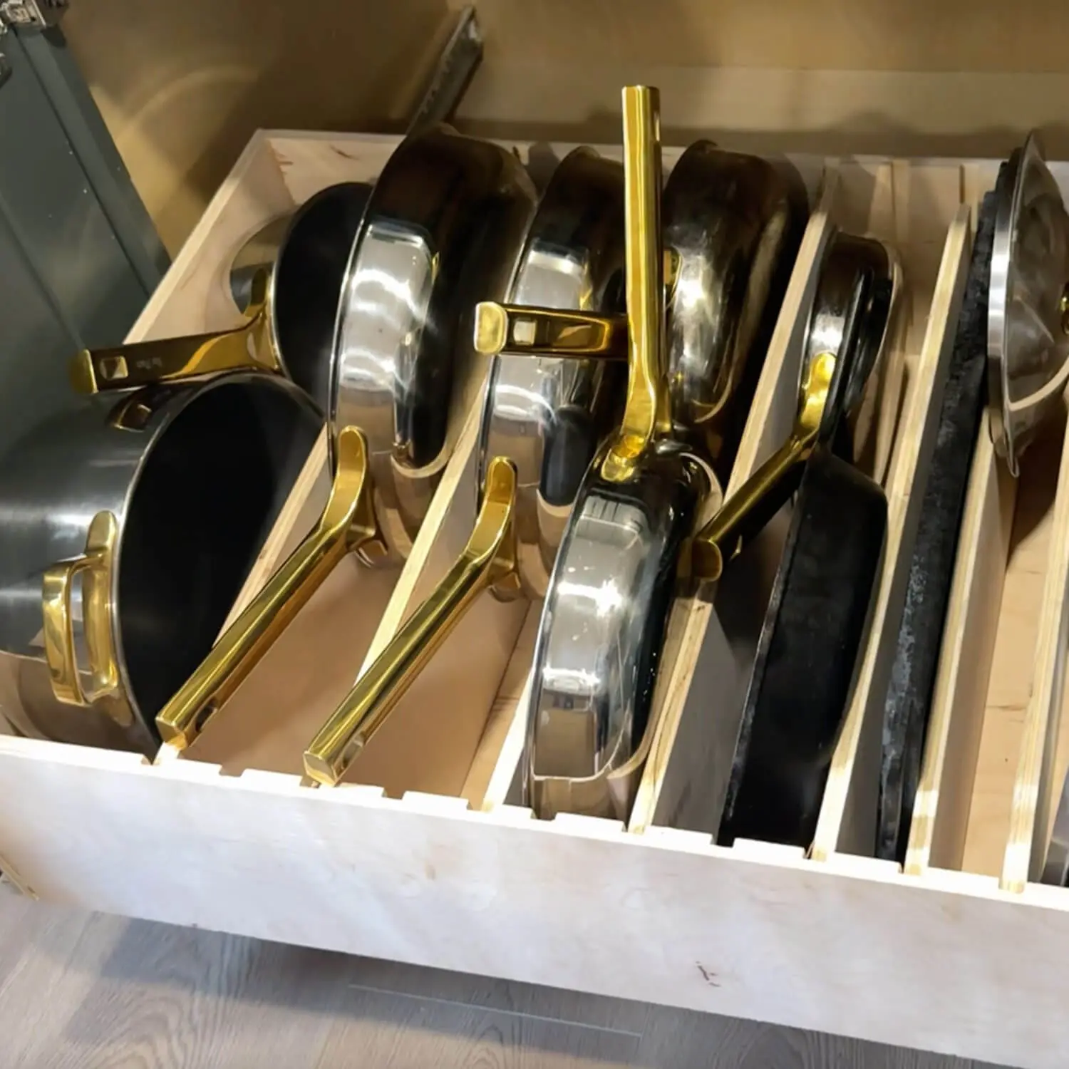 Close-up of a DIY slide-out cabinet drawer with vertical plywood dividers used as pots and pans storage, holding stainless steel cookware with brass handles standing upright in individual slots.