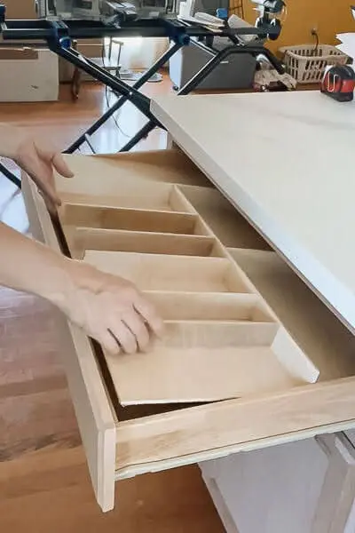 Hands sliding a custom plywood utensil drawer organizer with divided compartments into a shallow kitchen drawer for neatly sorted cooking tools.