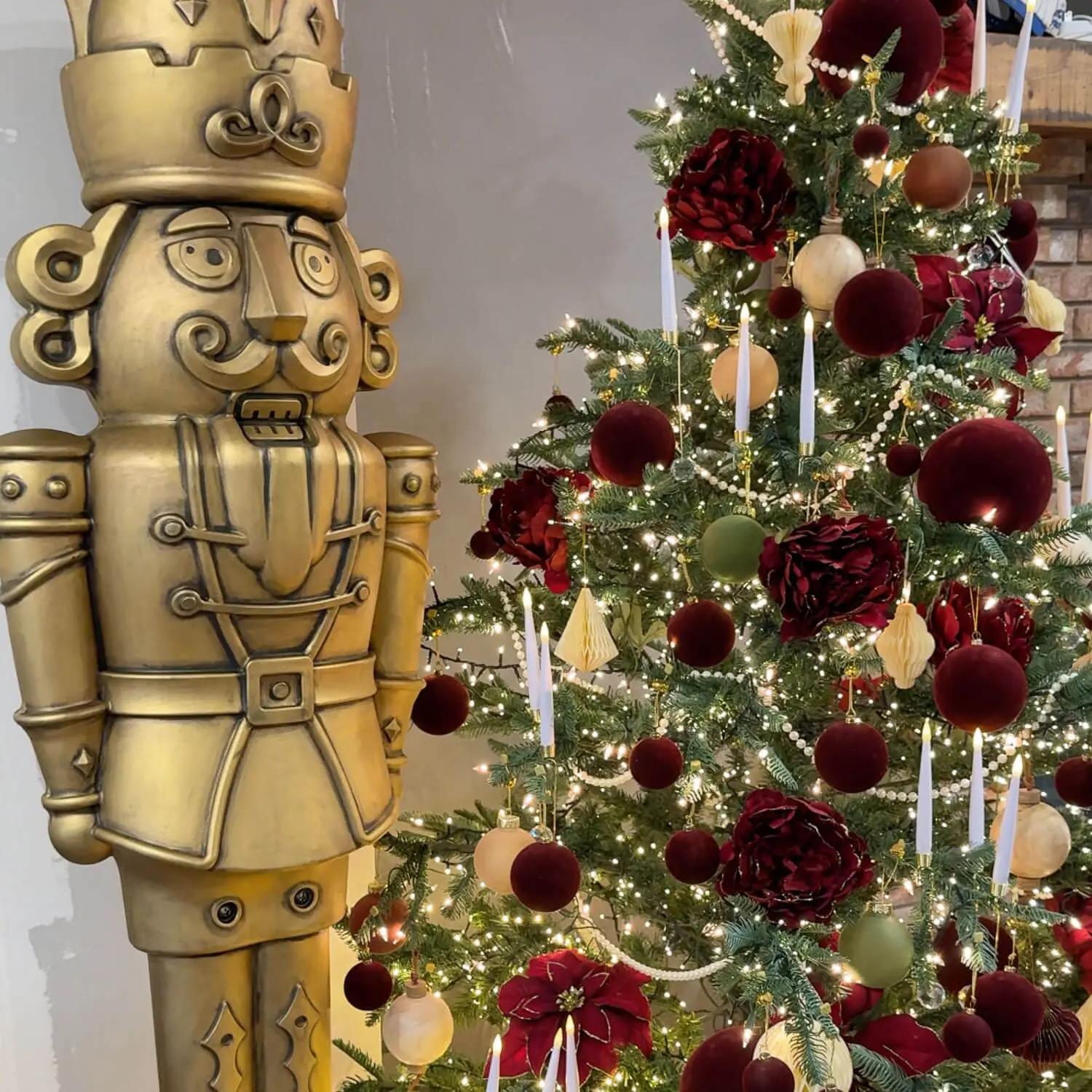 Antique bronze giant nutcracker standing beside a Christmas tree decorated with burgundy, green, and neutral ornaments and glowing candles.