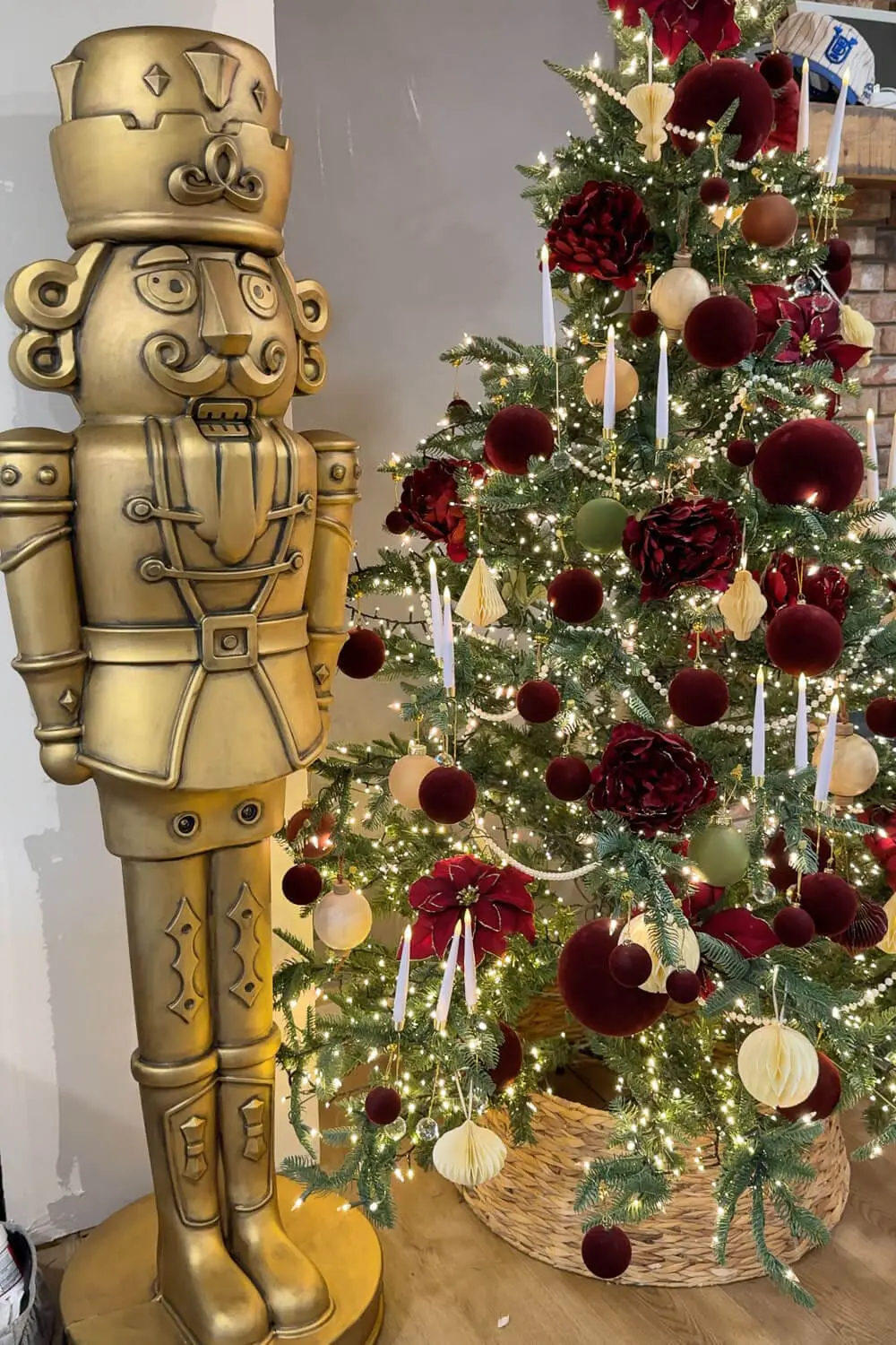 Full view of a tall antique bronze giant nutcracker next to a lit Christmas tree layered with burgundy ornaments, pearls, and candles.