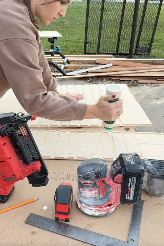 Woman applying wood glue to routed grooves on a plywood panel while building a DIY slide-out drawer for custom pots and pans storage, with nail gun and sander on the workbench.