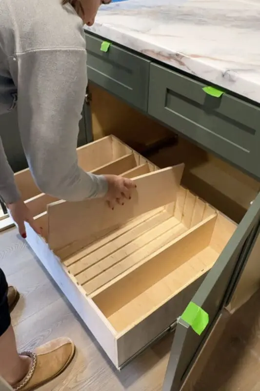 Woman sliding a tall plywood divider into the routed grooves of a slide-out drawer box, customizing the adjustable organizer for pots and pans storage in a lower kitchen cabinet.