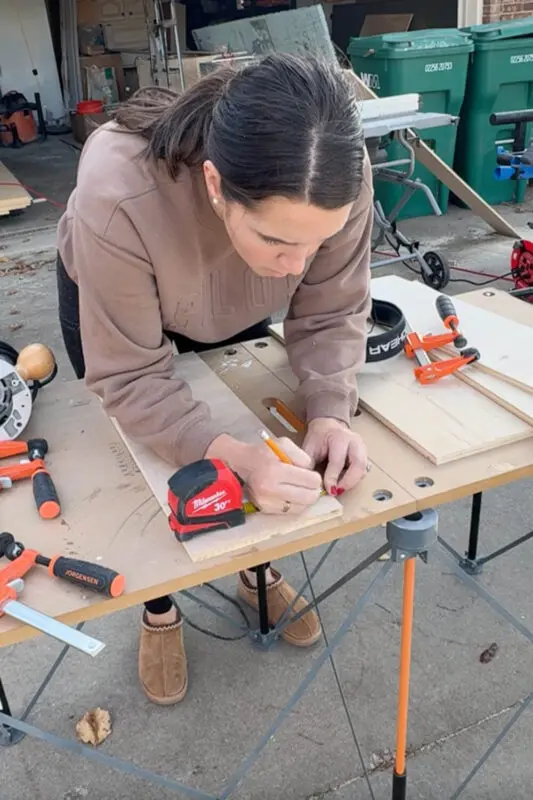 Woman marking measurements on a plywood board at an outdoor workbench, laying out where the grooves will be cut so the interlocking drawer pieces for the organizer slide together.