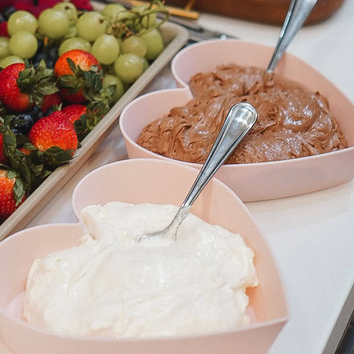 Heart-shaped bowls of marshmallow fluff fruit dip and chocolate fruit dip with spoons beside a fruit tray of strawberries, grapes, and blueberries.