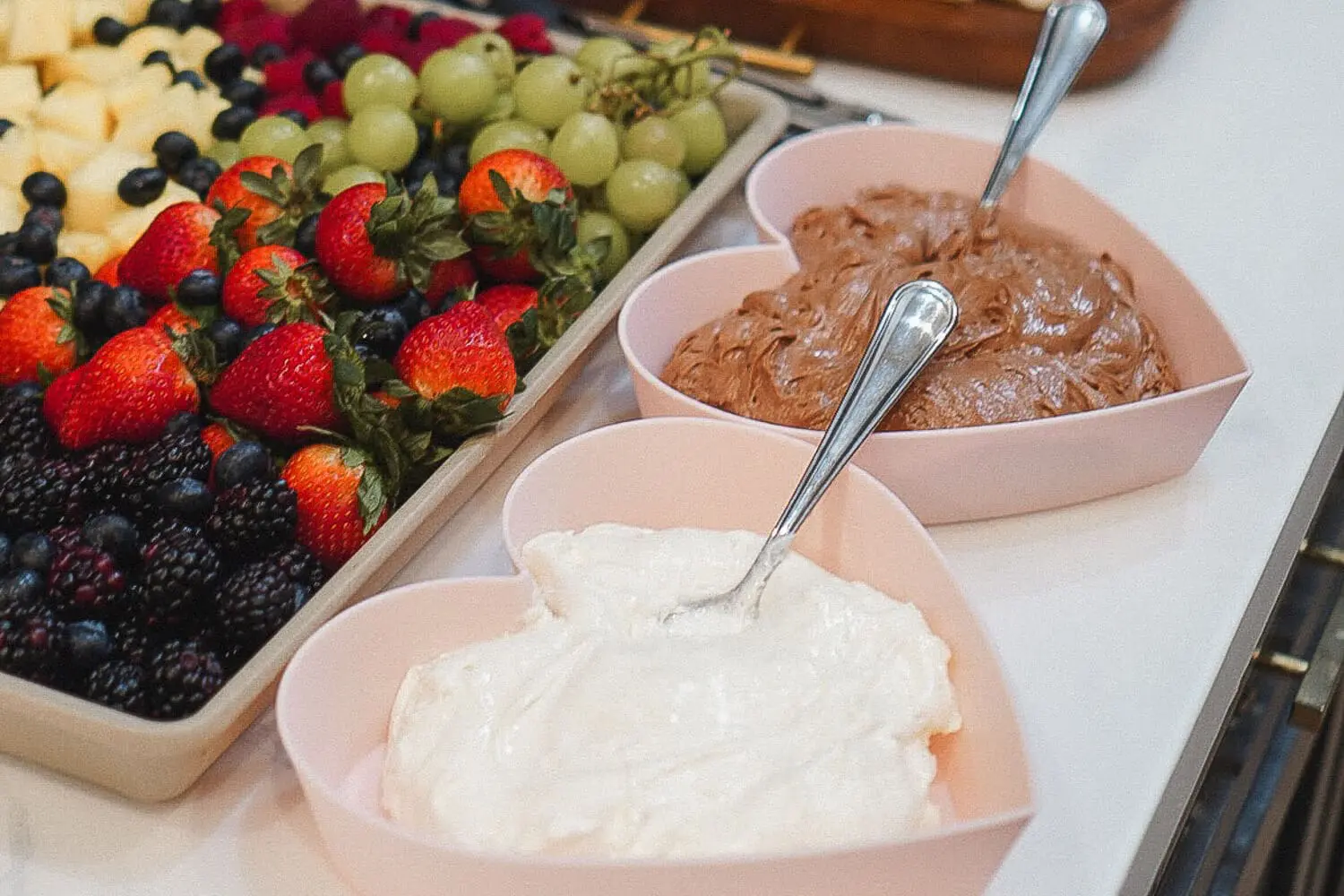 Heart-shaped bowls of marshmallow fluff fruit dip and chocolate fruit dip with spoons next to a colorful fruit tray of strawberries, grapes, blueberries, and blackberries.