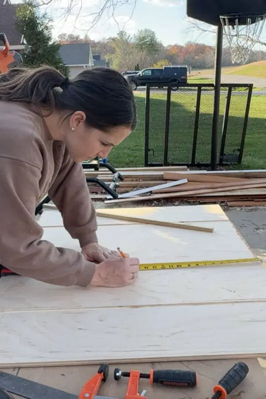 Woman measuring a large sheet of plywood with a tape measure and pencil, marking out the layout for a DIY slide-out drawer for pots and pans storage.