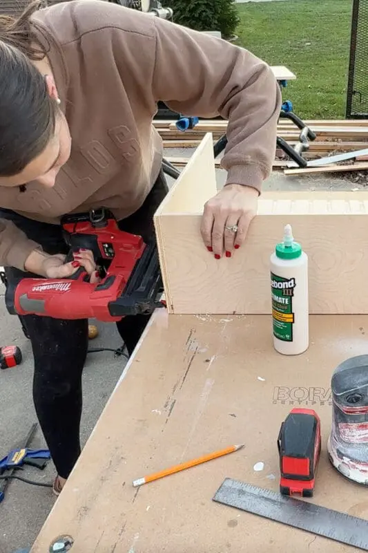 Woman using a cordless brad nailer to attach the side of a plywood drawer box, assembling a sturdy slide-out cabinet for organized pots and pans storage.