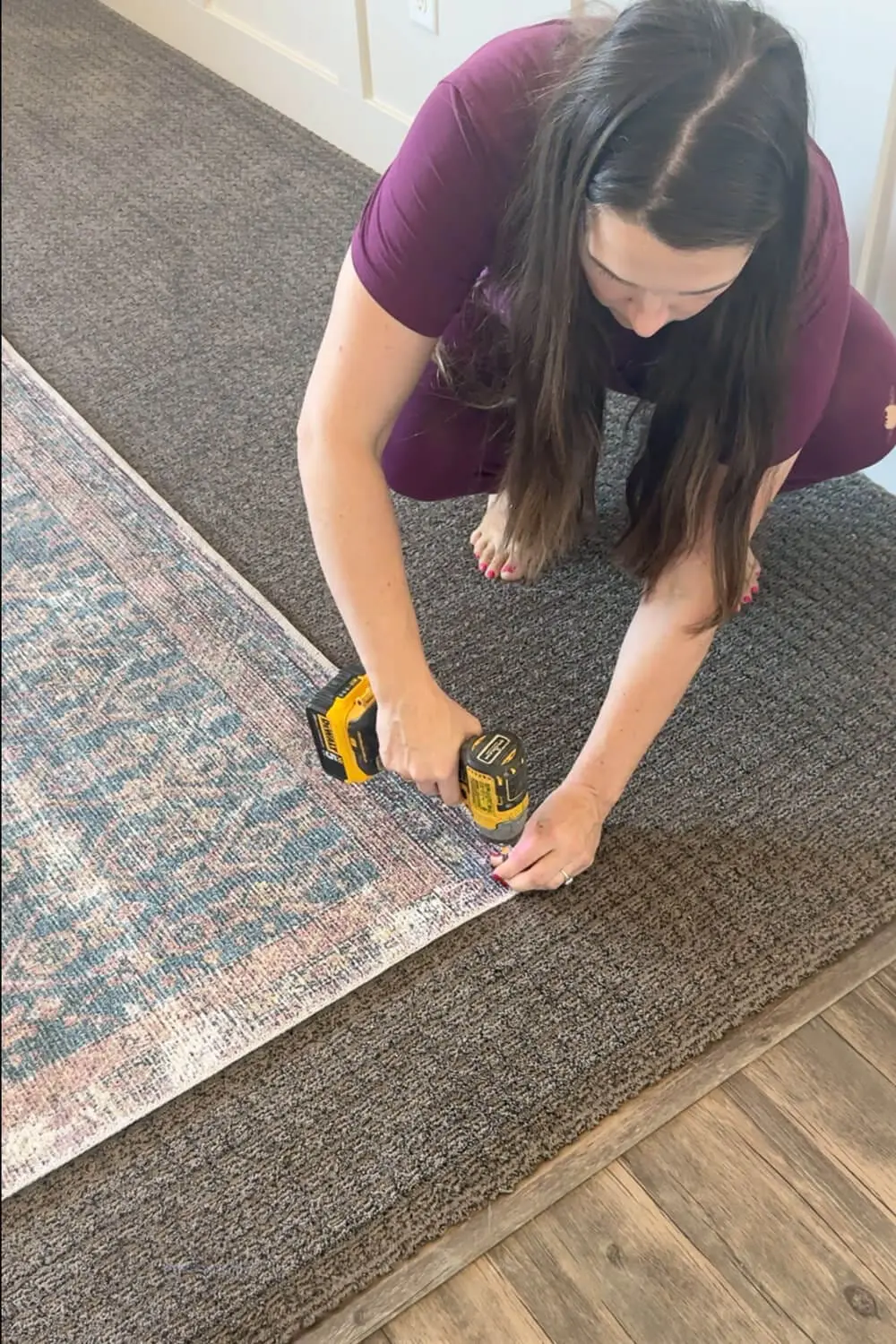 A woman using a drill to screw the corner of an area rug into the carpeted floor to stop sliding during a living room makeover. Works way better than rug tape.