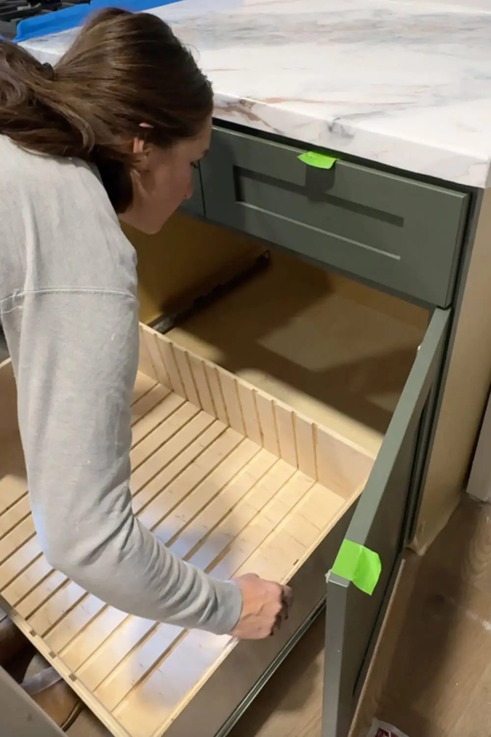 Woman is putting in an empty slide-out plywood drawer with routed grooves, testing the fit inside a lower kitchen cabinet before adding dividers for pots and pans storage.