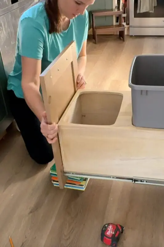 Carefully holding the cabinet door in place against the plywood double trash can drawer, using books to support the weight while installing a DIY pull out trash can cabinet.