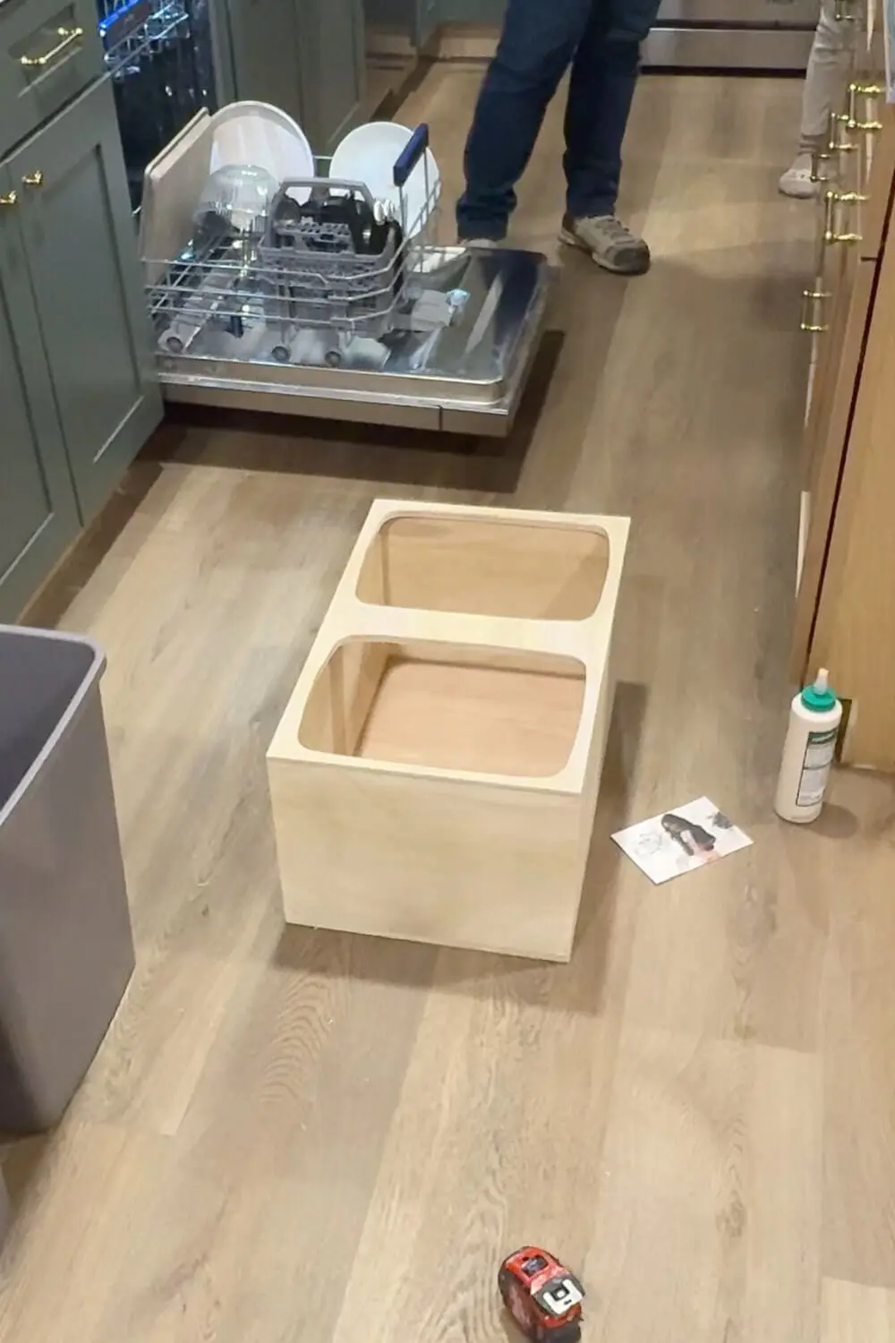 Unfinished plywood box with two rounded trash can openings sitting on the kitchen floor, showing the assembled insert for a DIY pull out double trash can cabinet.