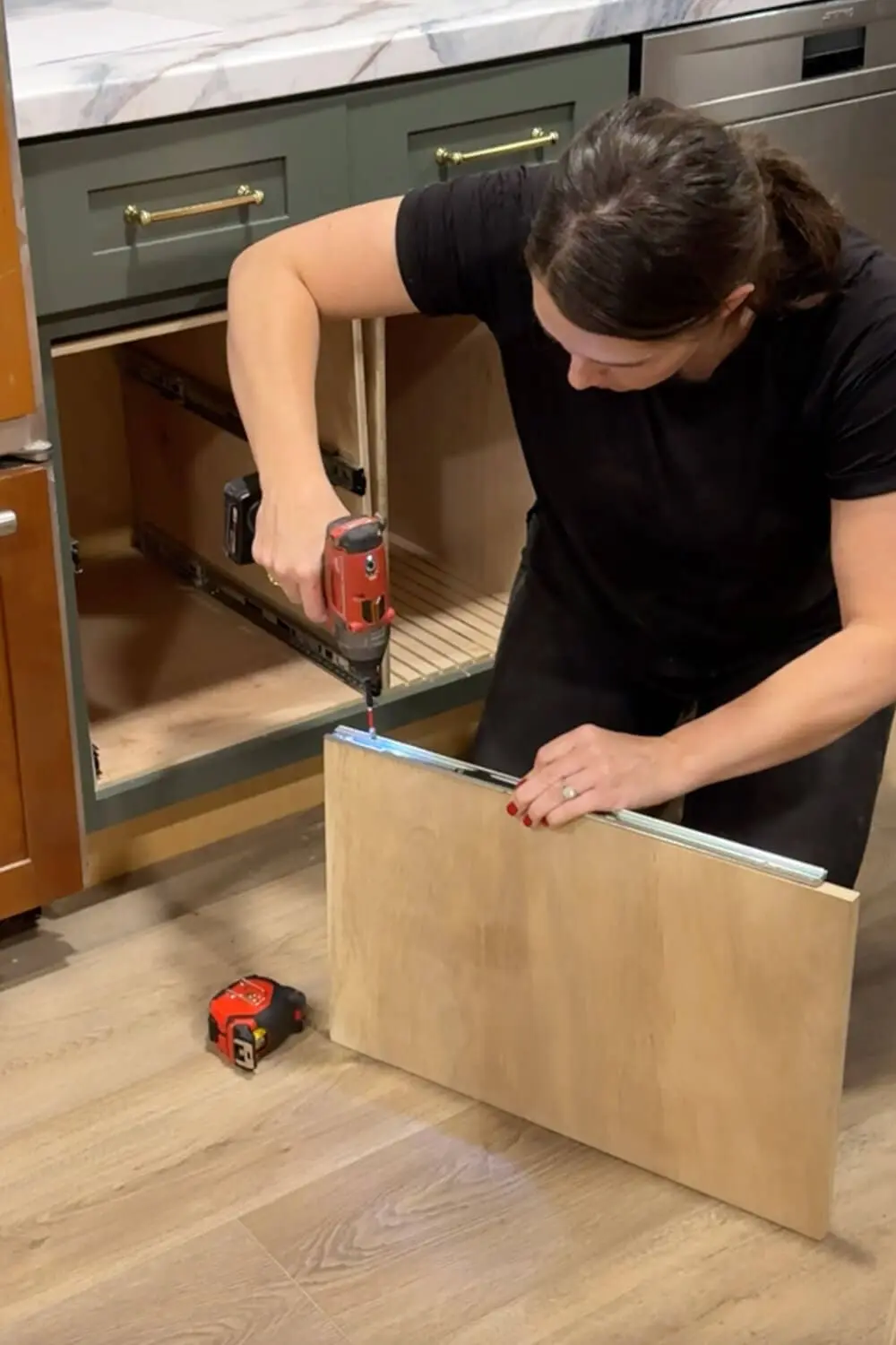 Woman using a drill to attach a drawer slide to the edge of a plywood shelf panel while building a DIY pull-out shelf for organizing food storage containers in a lower kitchen cabinet.