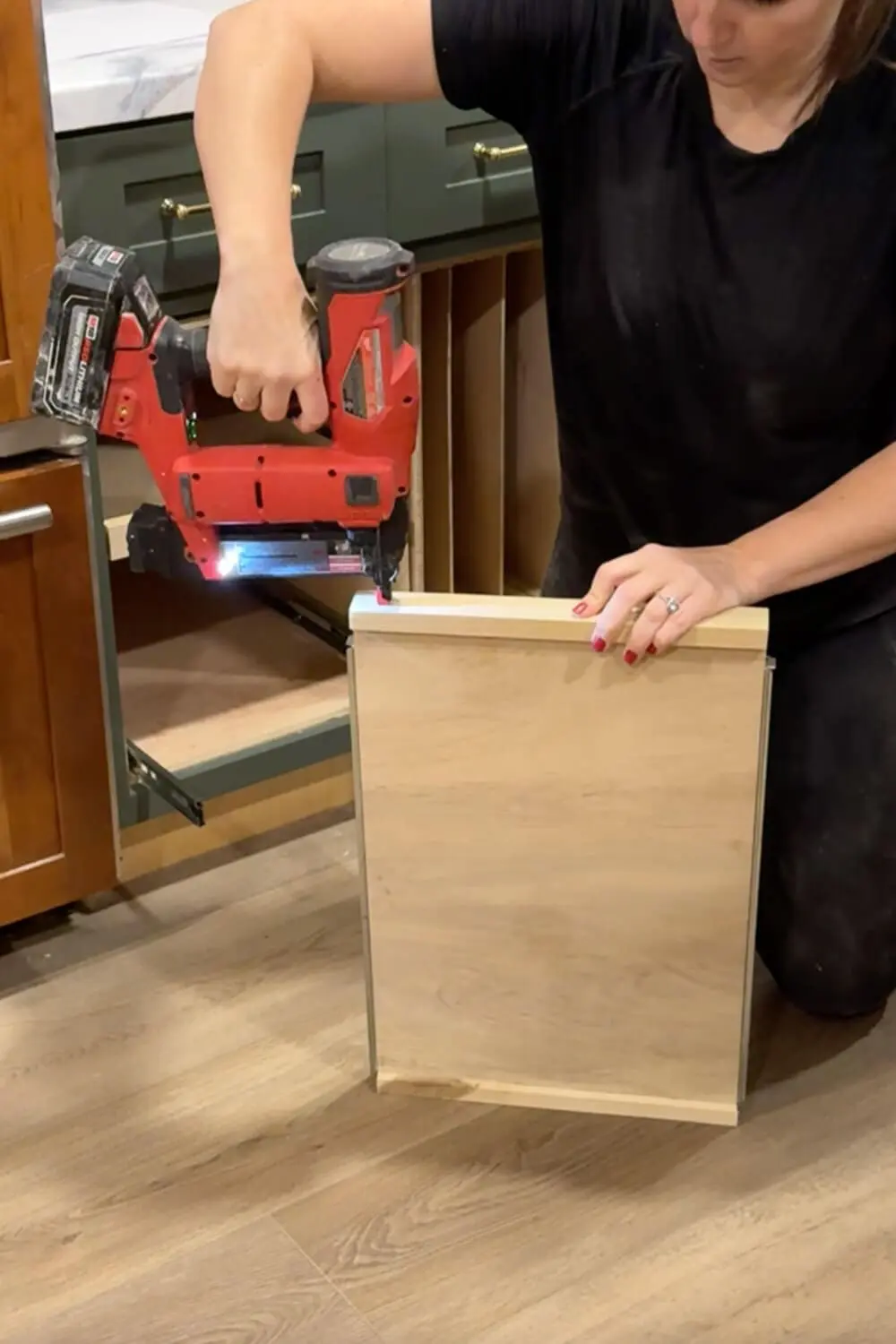 Woman using a cordless brad nailer to attach a small front rail to a plywood pull-out shelf for organizing food storage container lids in a lower kitchen cabinet.