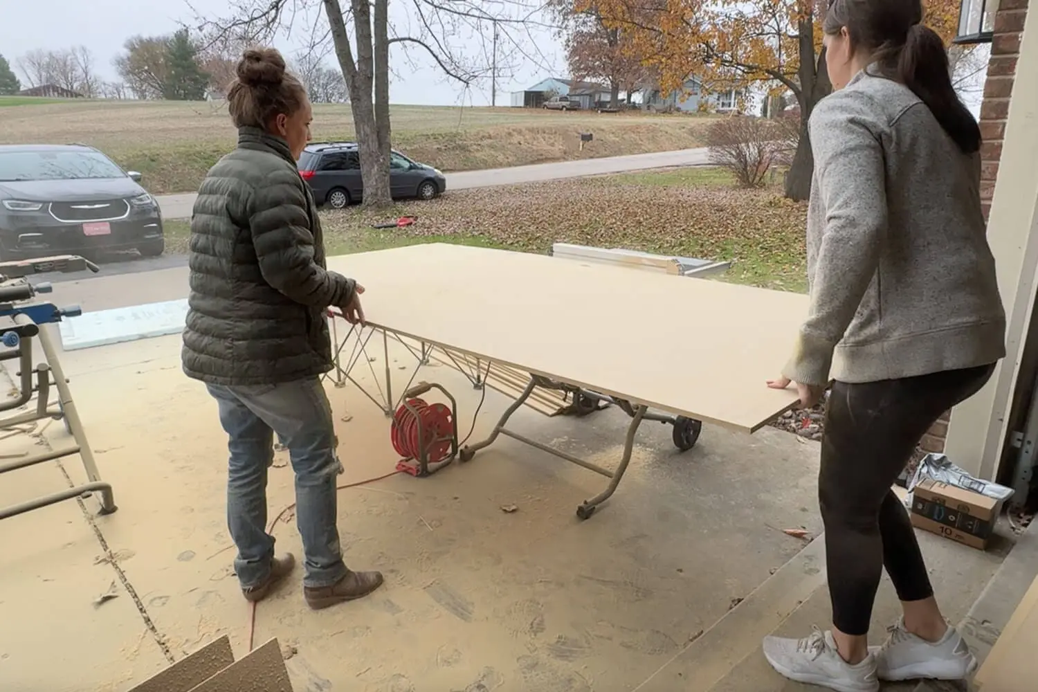 Two women feeding a large sheet of plywood through a table saw in the garage to cut pieces for a DIY vertical storage cabinet insert