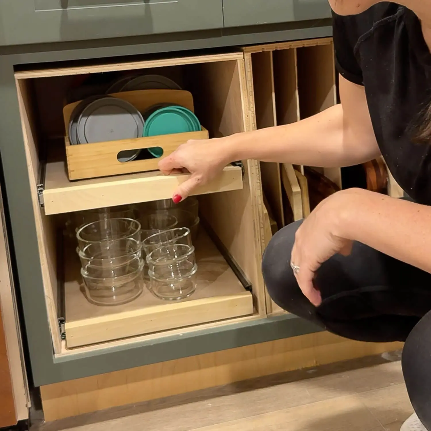 Close up of a DIY plywood drawer organizer holding glass food storage containers on the bottom pull-out shelf and a wooden bin of Tupperware-style lids on the upper slide-out shelf.
