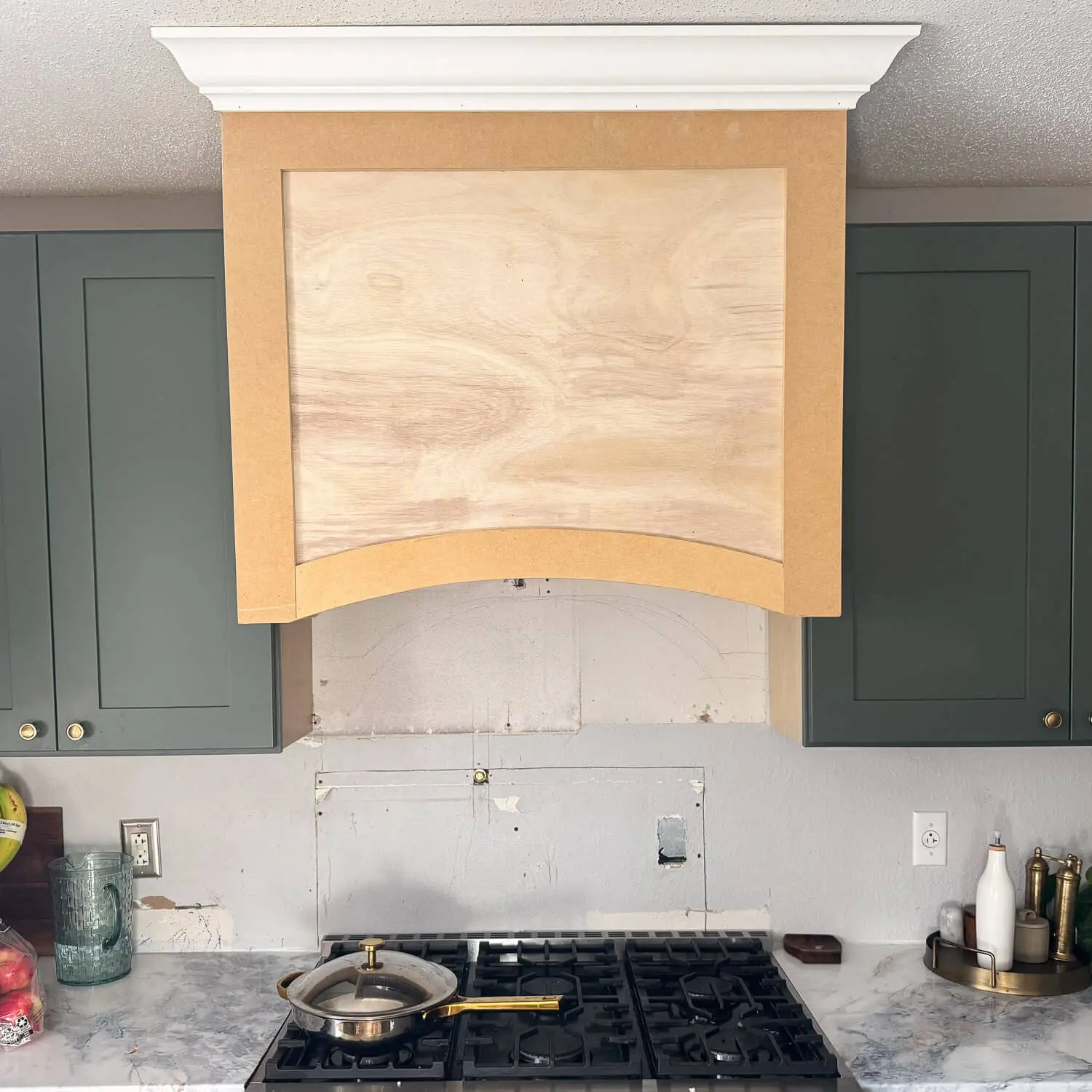 Closer look at a DIY wood range hood cover build with a plywood face panel and arched lower detail, framed in with MDF and topped with white crown molding, installed between dark green cabinets during a kitchen remodel.