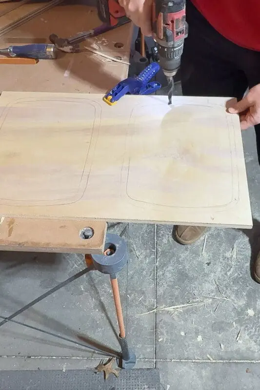 Drilling starter holes inside the traced trash can openings on a plywood panel, prepping to cut out the bin openings for a DIY pull out double trash can cabinet.